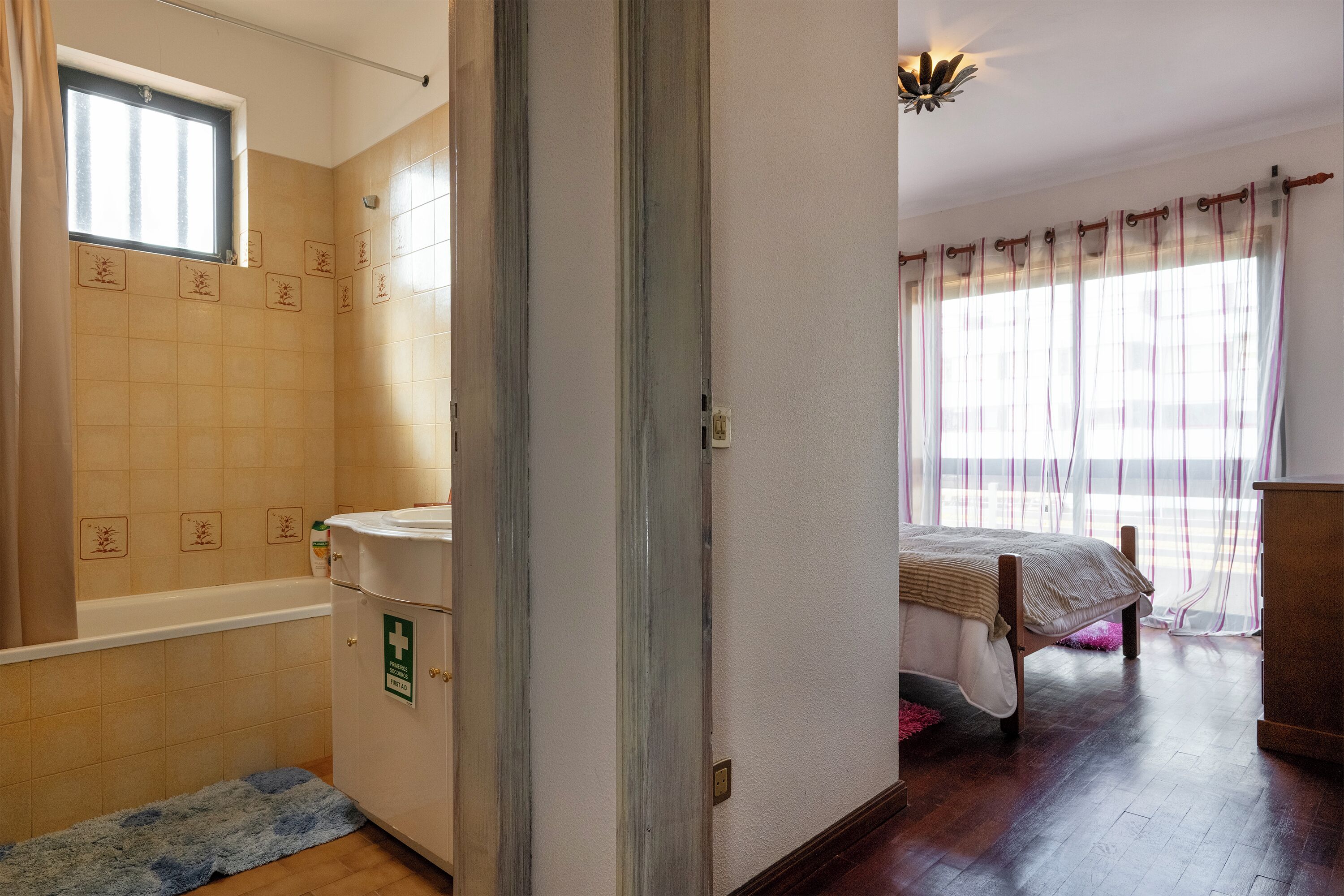 Ein Schlafzimmer mit einem Bett, einem Fenster mit Vorhängen, Holzboden und einem Badezimmer mit Badewanne und Waschbecken.