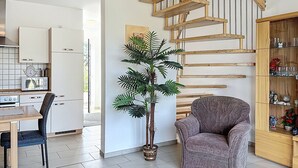 Una cocina moderna con armarios blancos, un comedor con mesa y sillas, una planta en maceta, un sillón gris y una escalera de madera.