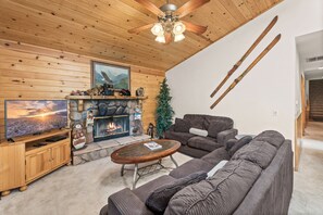 A cozy living room with a fireplace, a large flat-screen TV, a ceiling fan, and a ski rack. - Spacious Log Cabin w/ Game Room & AC Near Bear Mountain (Big Bear City)