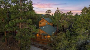 A two-story cabin surrounded by dense forest, with a visible deck and a chimney. - Spacious Moonridge Log Cabin with A/C, Game Room, 2 Fireplaces & Fenced Yard (Big Bear City)
