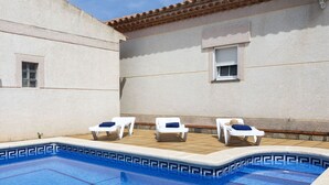 Ein Schwimmbecken mit vier Liegestühlen, einer gefliesten Terrasse und einem Gebäude mit einem gefliesten Dach und Fenstern.