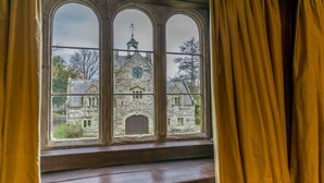 Blick durch ein dreigeteiltes Fenster auf ein Steinhaus mit einer Uhr und Wetterfahne.
