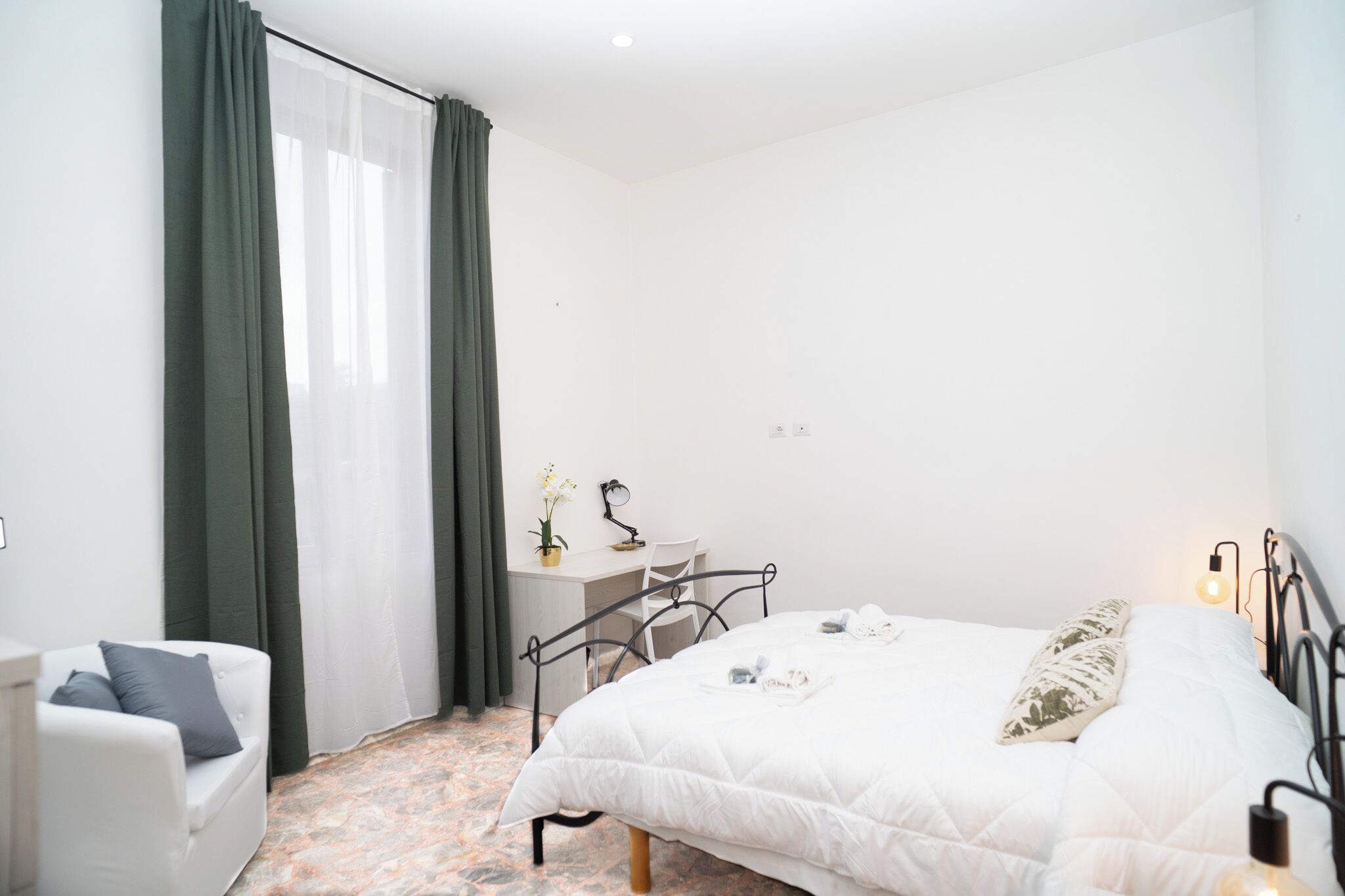 A bedroom with a bed, a desk, a chair, and a small table with a lamp.