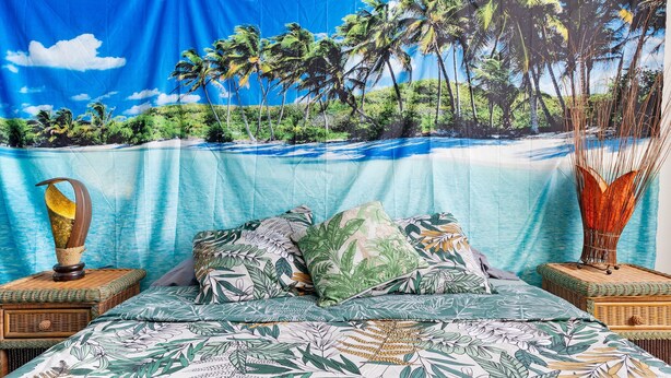 Een bed met een tropisch strandtapijt als achtergrond, een houten nachtkastje met een lamp en een geweven mand.