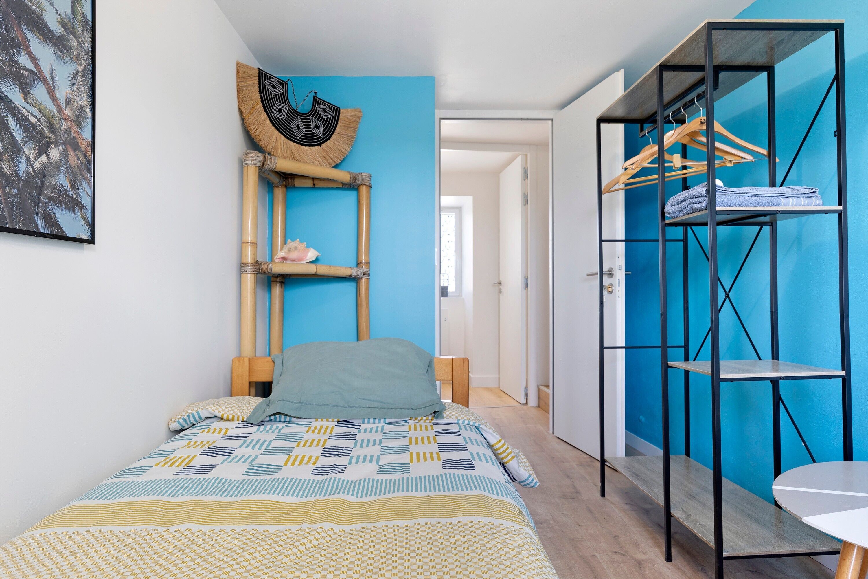 A bedroom with a bed, a wall-mounted shelf with clothes, a door, and a view into another room.