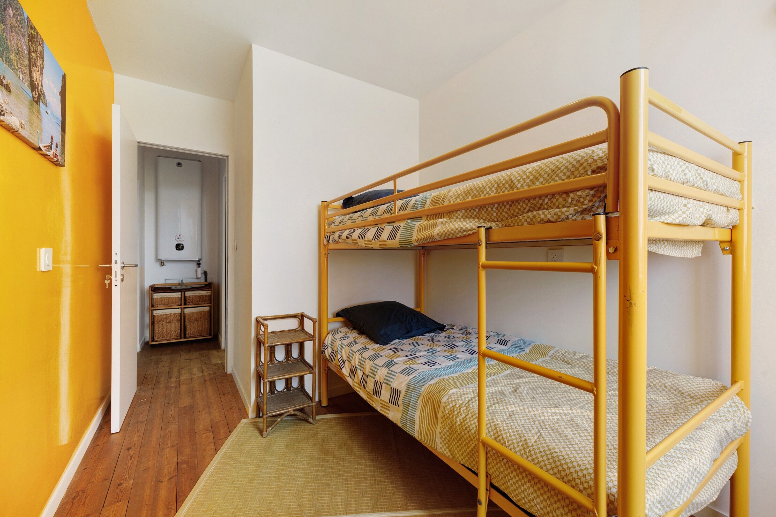 A bunk bed room with a wooden floor, a small stool, and a door leading to a kitchen area.