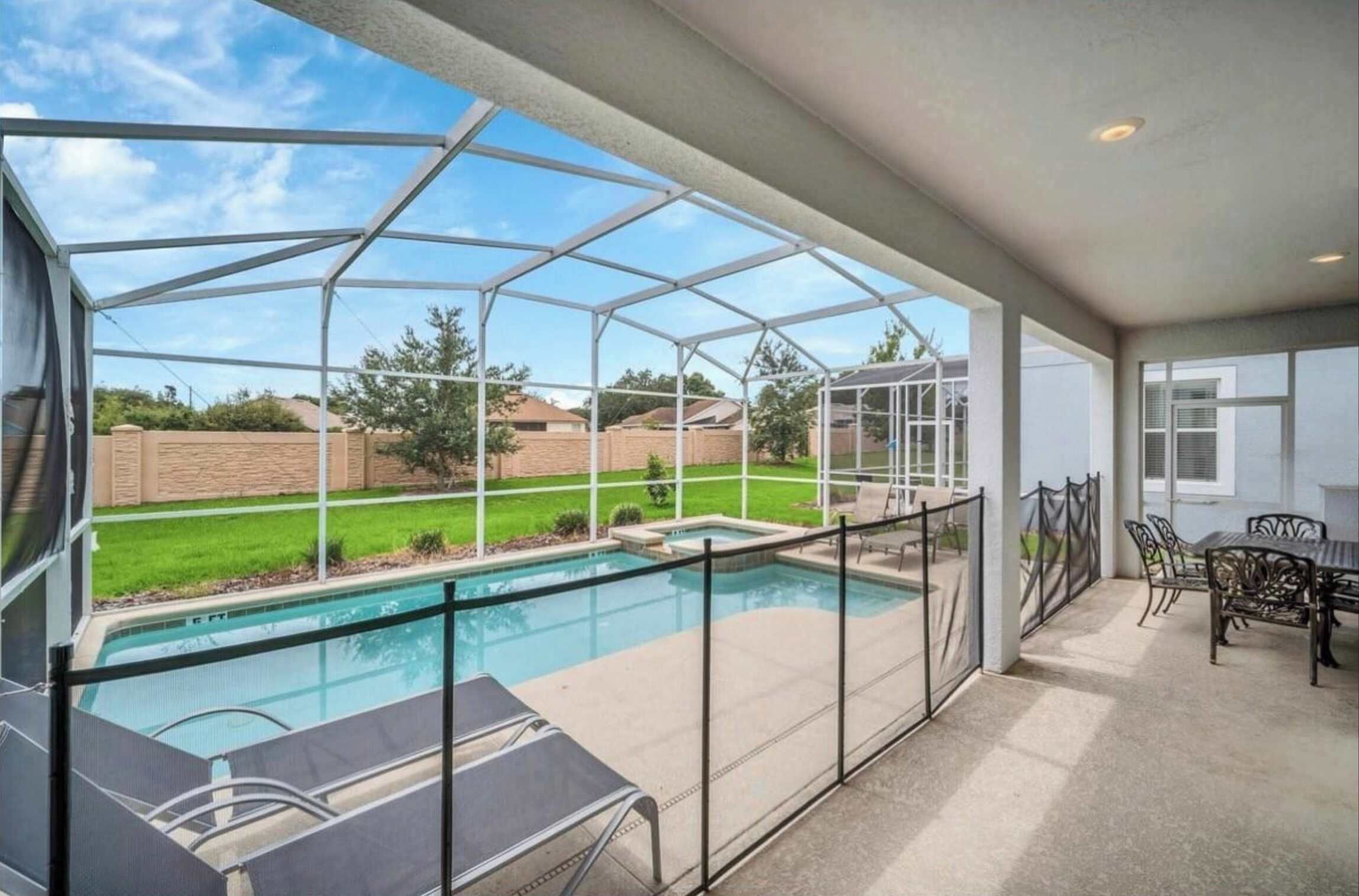 A covered outdoor swimming pool area with a glass enclosure, a patio with a table and chairs, and a view of a residential neighborhood.
