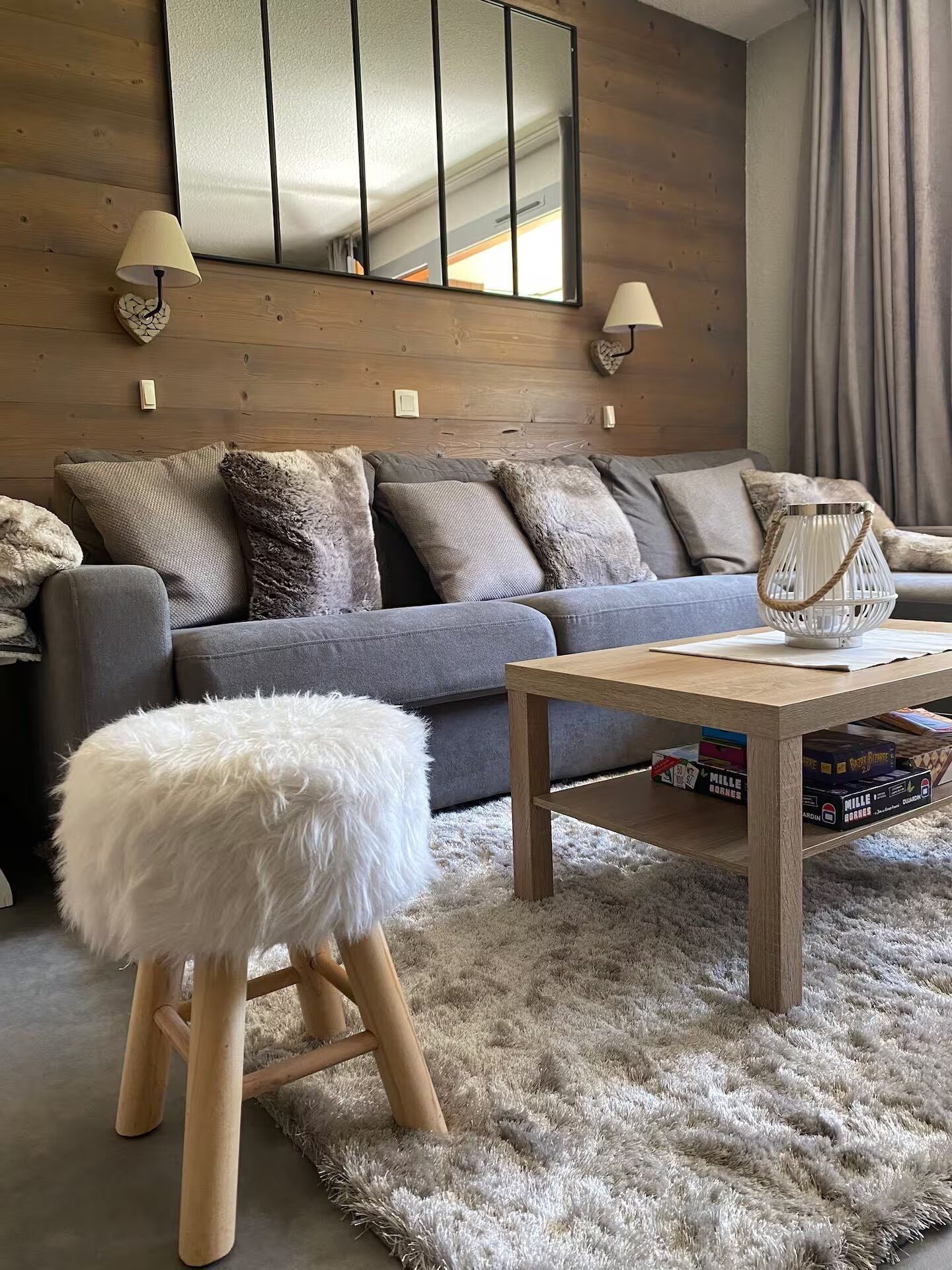 Una sala moderna con un sofá gris, un pouf blanco mullido, una mesa de centro de madera y un espejo en la pared superior.