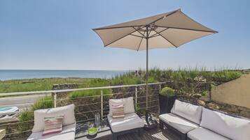A deck with white cushioned seating, a large beige umbrella, and a view of the sea.
