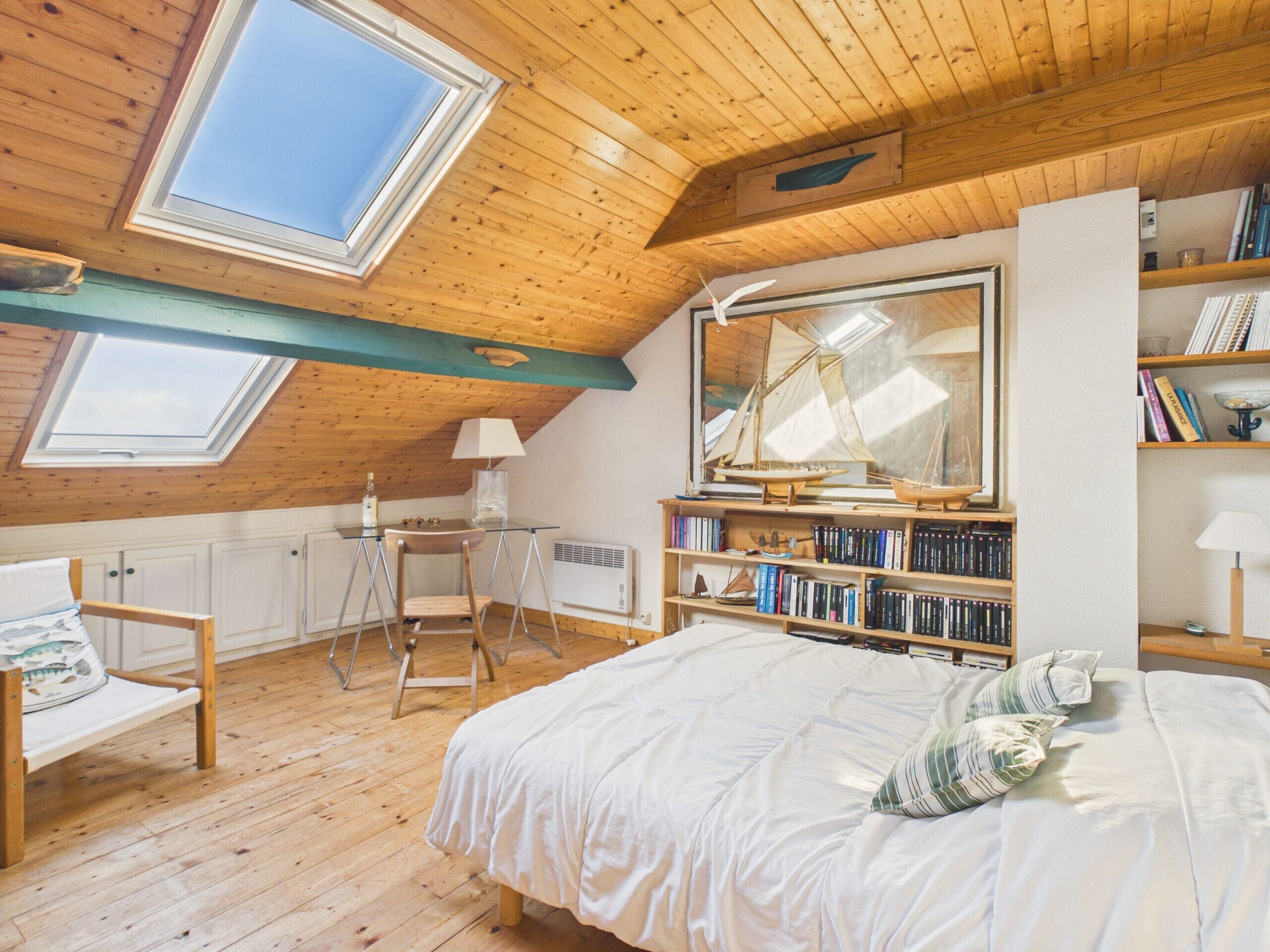 Un dormitorio acogedor en el desván, con techo inclinado, vigas de madera, tragaluces, una cama, un escritorio y una estantería.