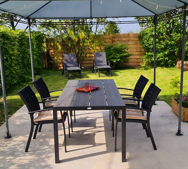 A covered patio with a table and chairs, surrounded by greenery.