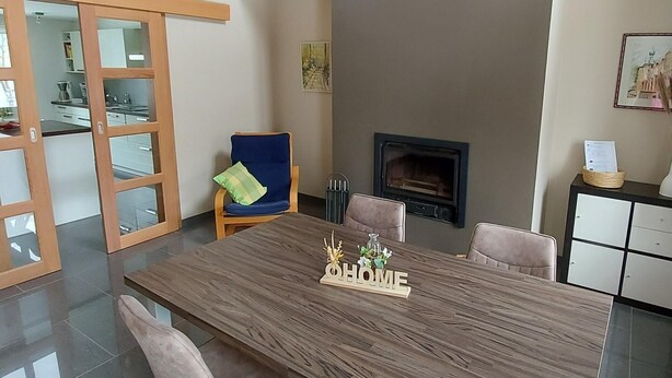 A modern dining area with a large wooden table, cushioned chairs, a fireplace, and a view of the kitchen through glass doors. - Cozy House for 4 in the Heart of the French Ardennes – Laundry, Bike Storage, Near Rocroi & Belgium (Gué-d'Hossus)