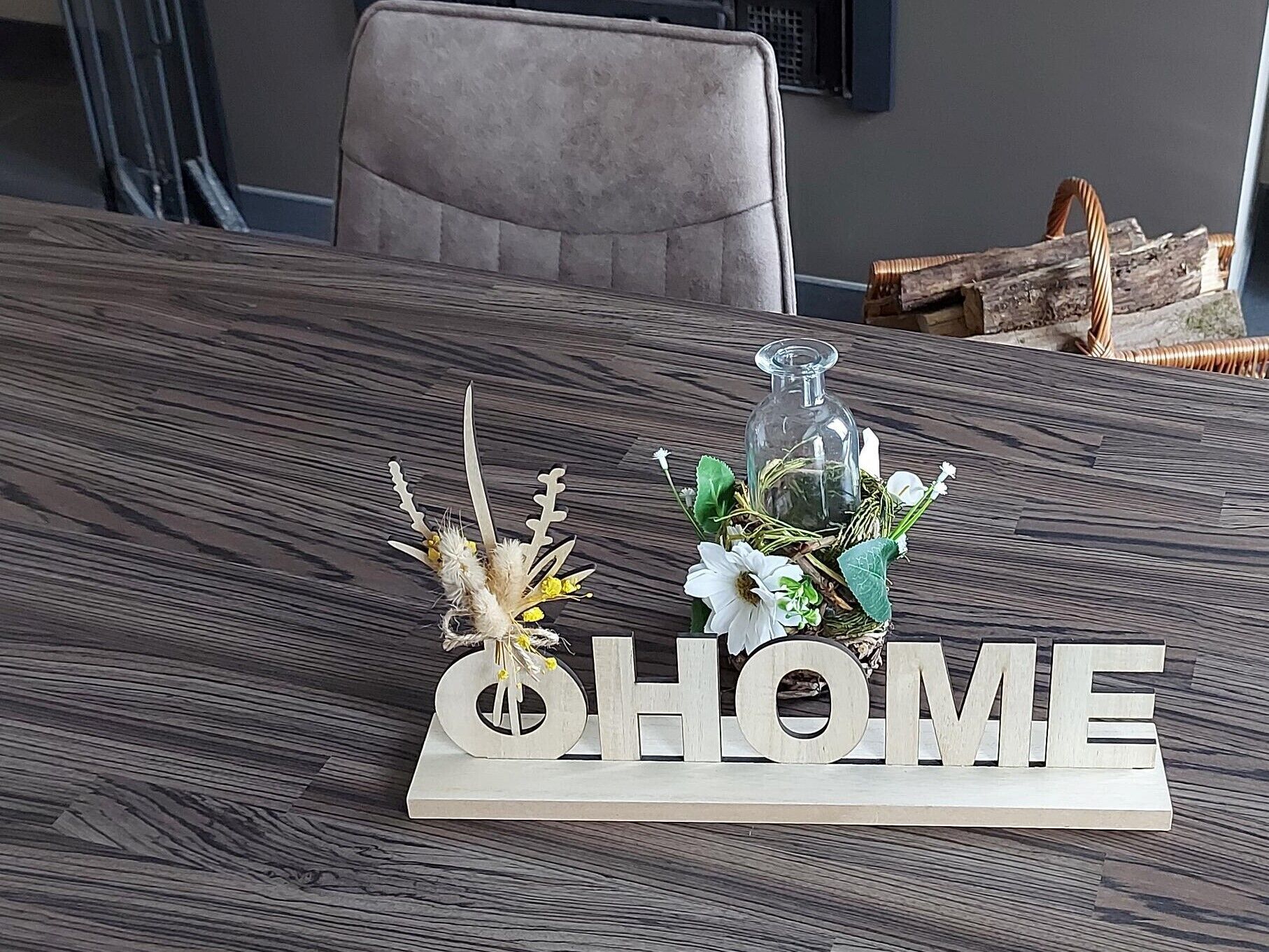 Une table à manger agrémentée d’un élément décoratif central composé d’un vase en verre, de fleurs et d’une petite plaque en bois sur laquelle est inscrit le mot « MAISON ».