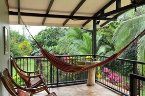 Un balcon agrémenté d’un hamac et de chaises en bois, entouré d’une végétation luxuriante et doté d’un sol carrelé.