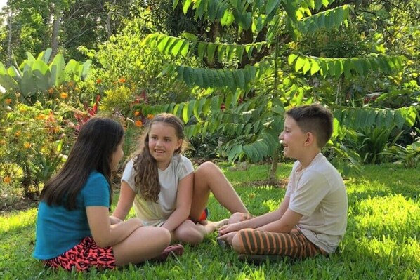 Three people sitting on grass in a garden with various plants and trees in the background.