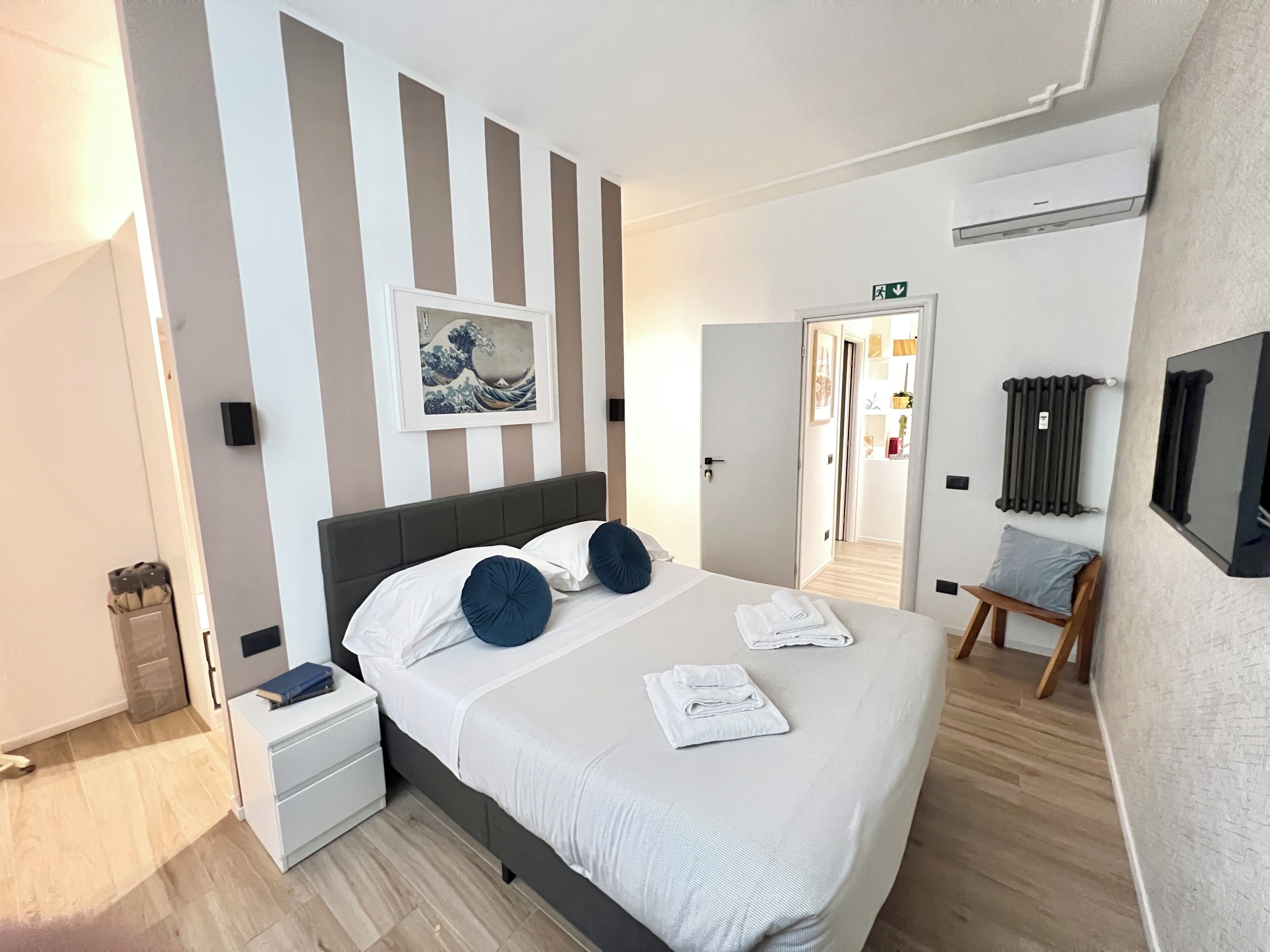 Hotellihuone, jossa on sänky, yöpöytä, tuoli, patteri ja taulu seinällä.