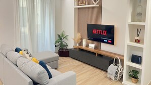 A modern living room with a grey sofa, a flat-screen TV mounted on the wall, and a wooden shelf with decorative items. - House Of Victories (Roma)