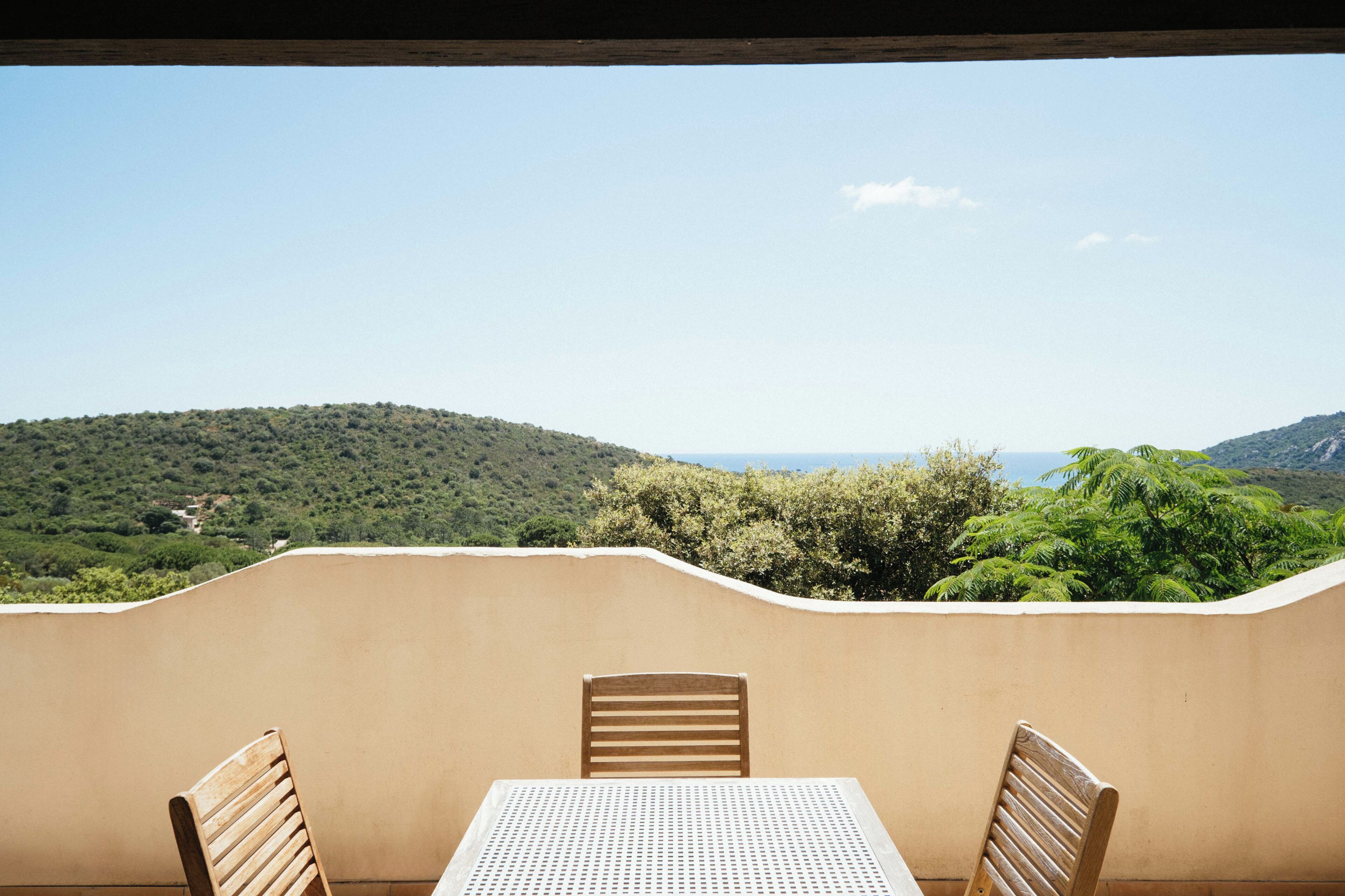 Un balcon avec une table et des chaises, donnant sur un paysage vallonné parsemé d’arbres et sous un ciel dégagé.