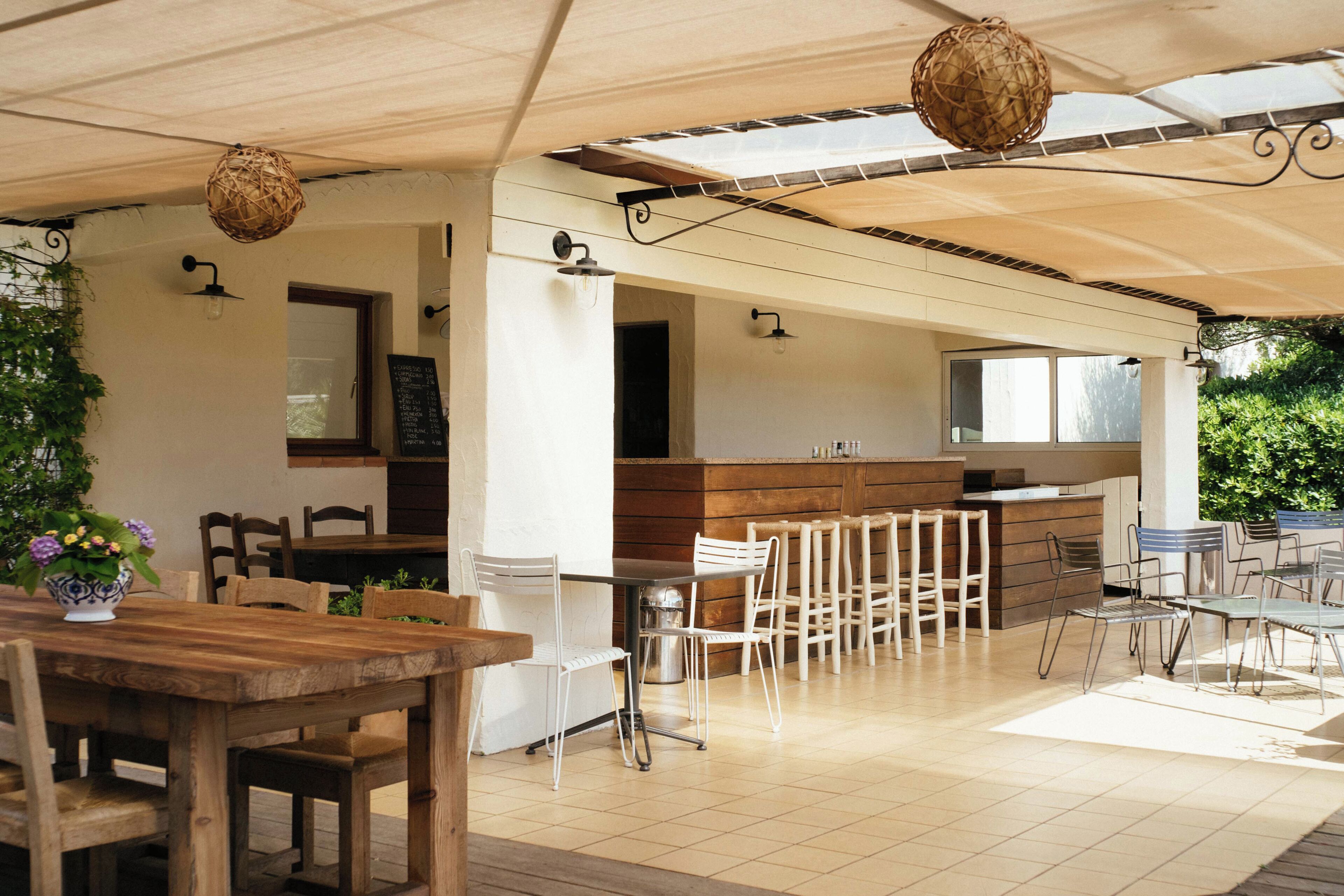 A covered outdoor dining area with wooden tables and chairs, a kitchen area with a bar, and hanging decorative light fixtures.