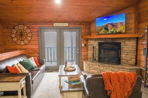 Ein gemütliches Blockhaushaus-Interieur mit einem Ledersofa, einem Kamin aus Naturstein, einem Fernseher, der über dem Kamin angebracht ist, und einer Holzbalkendecke.