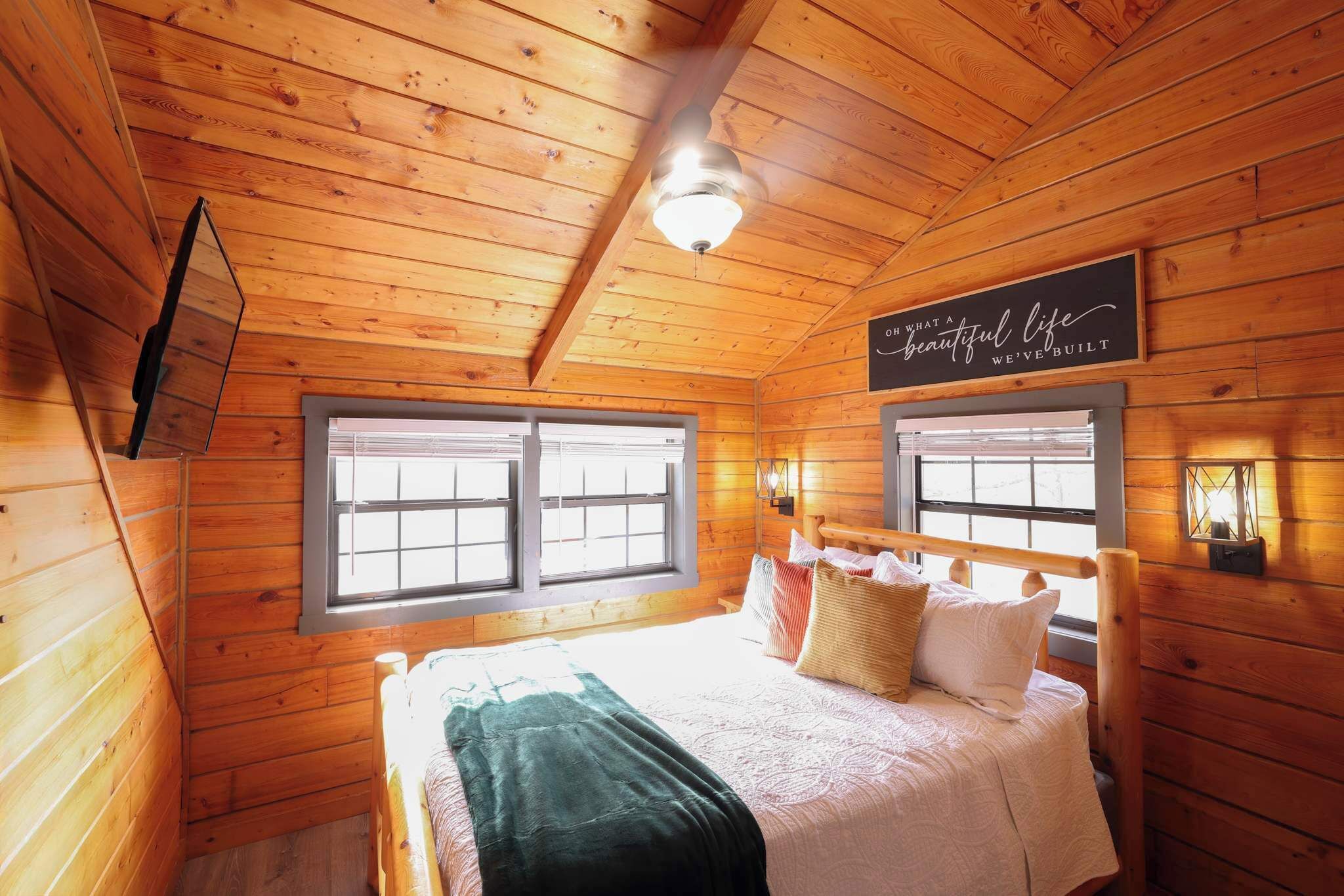 A cozy wooden cabin bedroom with a bed, two windows, a ceiling fan, and a sign on the wall.