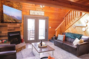 A cozy cabin interior with a stone fireplace, a television mounted on the wall, a wooden staircase, and a sign welcoming guests. - Hero's Peak | Theater, Arcade & Hot Tub (Sevierville)