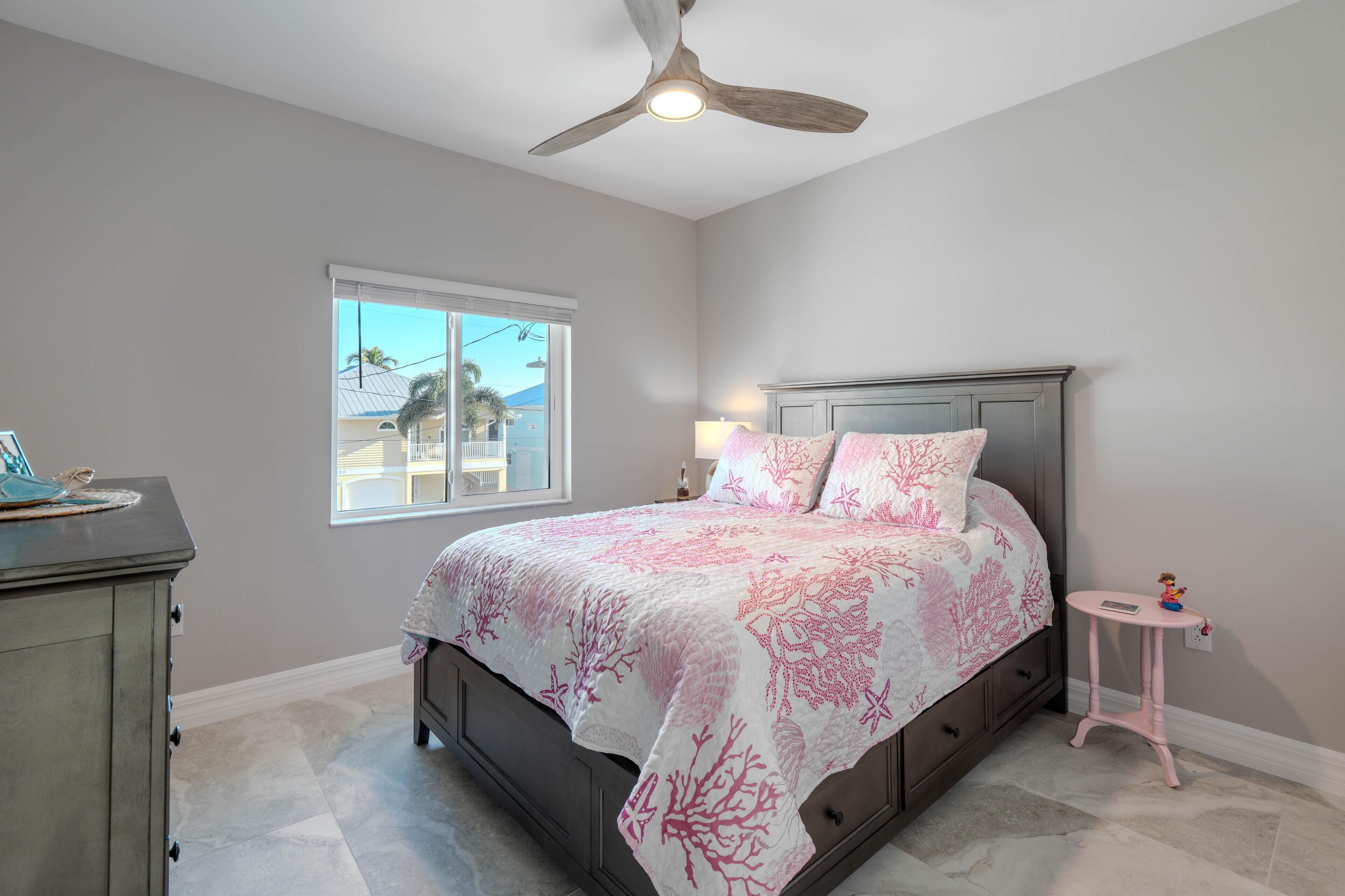 Une chambre à coucher comprenant un lit, une commode, une table de chevet, un ventilateur de plafond et une fenêtre donnant sur des palmiers.