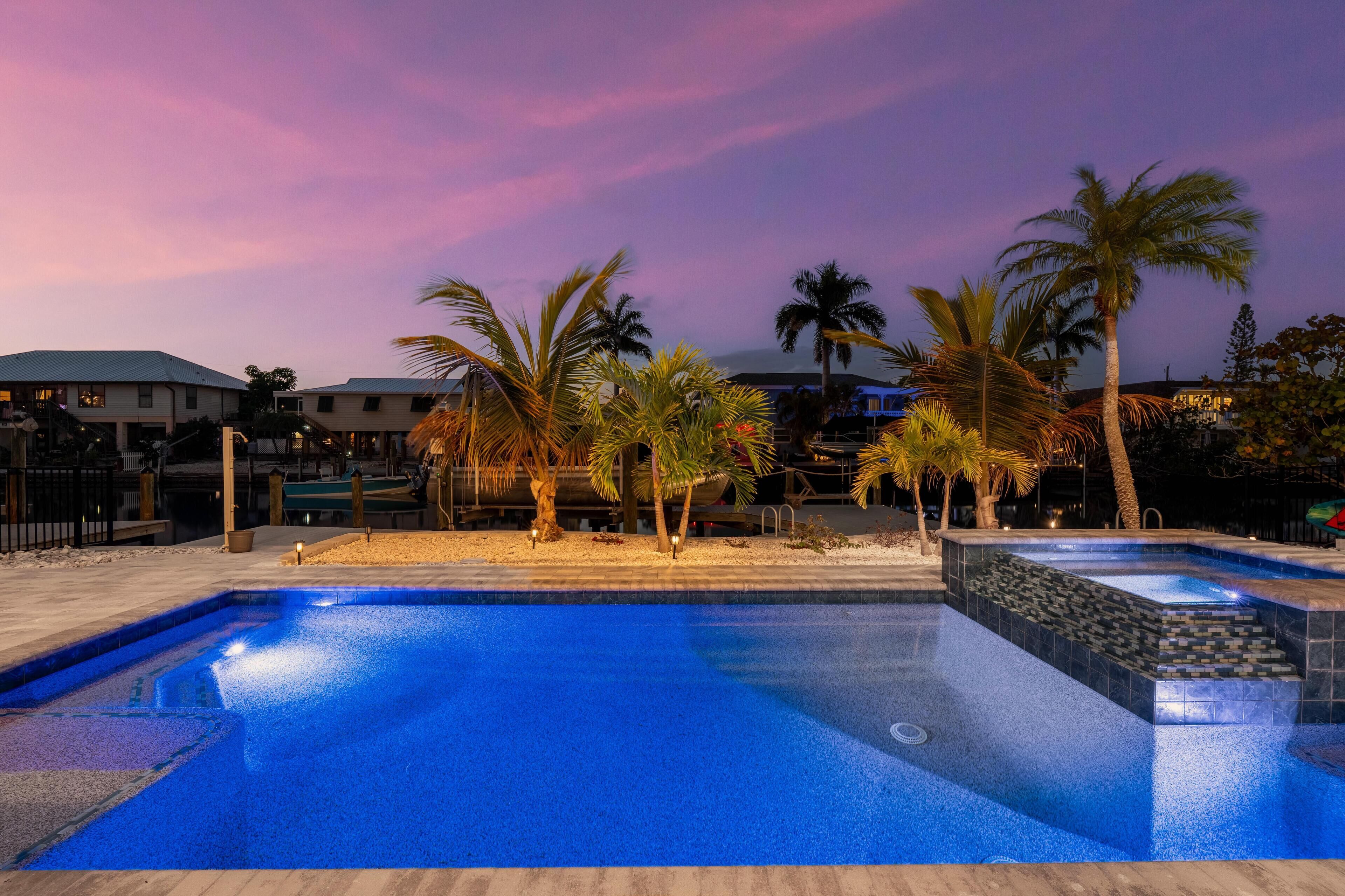 Une piscine dont le bord est recouvert de carreaux, entourée de palmiers et surplombée par un ciel au coucher du soleil.