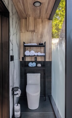 Une salle de bain moderne avec un mur en marbre, un lambris en bois, une étagère noire avec des serviettes, des toilettes et une poubelle.