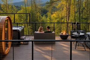 Un couple se détend sur une terrasse équipée d’un jacuzzi, de coins salon extérieurs et d’un foyer, avec une vue imprenable sur un paysage boisé.