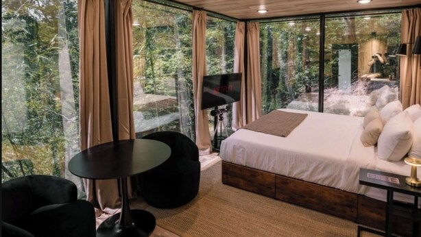 Une chambre moderne avec un grand lit, une petite table et une chaise. Un téléviseur est posé sur un trépied devant une fenêtre.