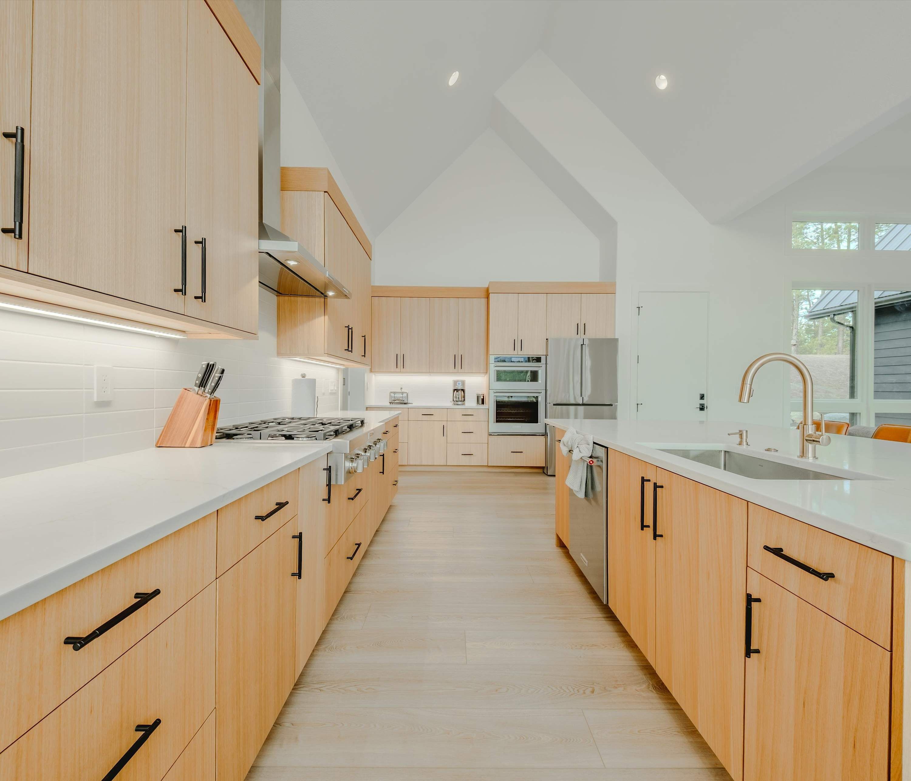 Een moderne keuken met houten kastjes, een wit aanrechtblad, roestvrijstalen apparatuur en een groot raam dat voor natuurlijk licht zorgt.