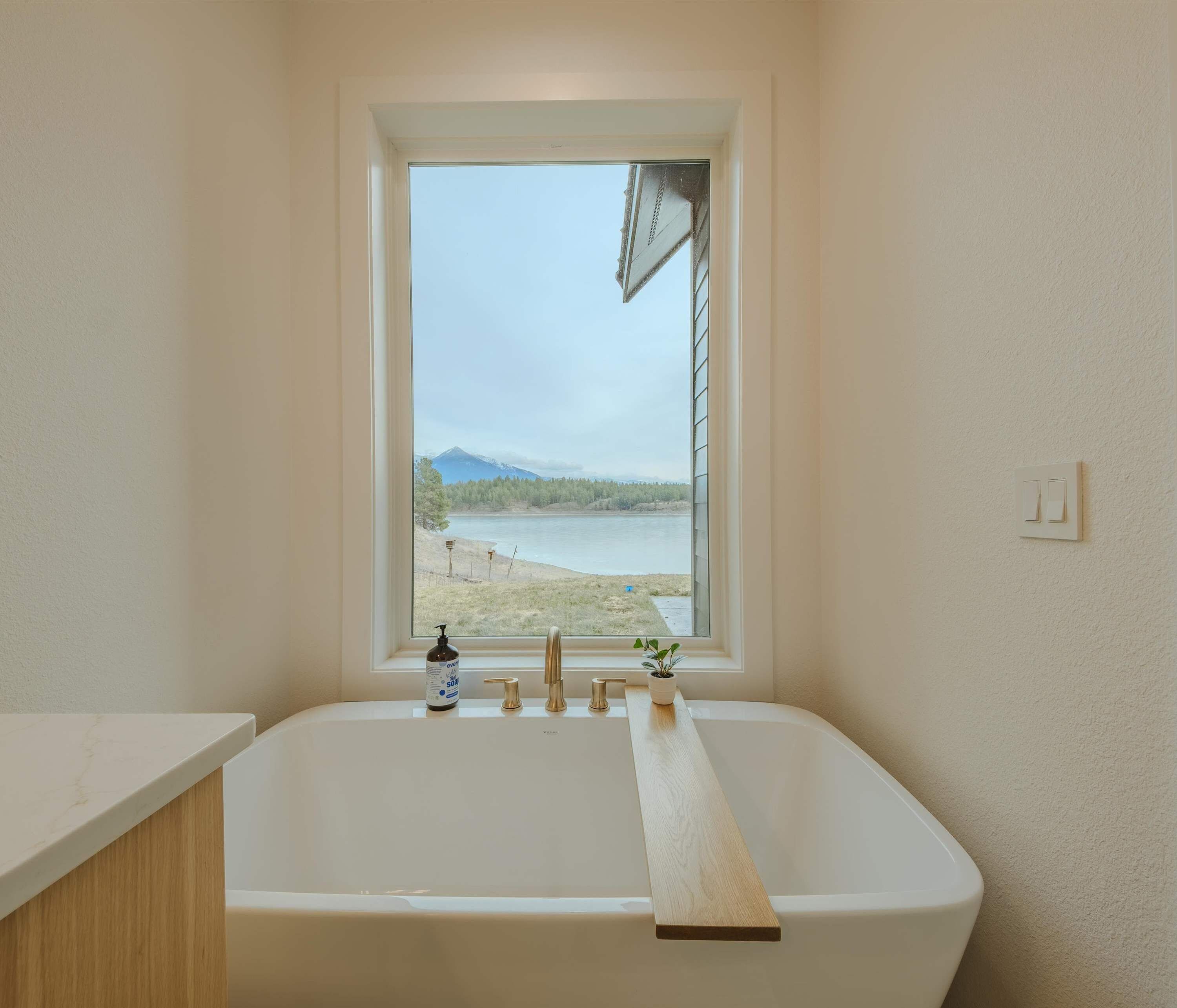 A bathroom with a bathtub, a window with a view of a lake and mountains, and a wooden shelf.
