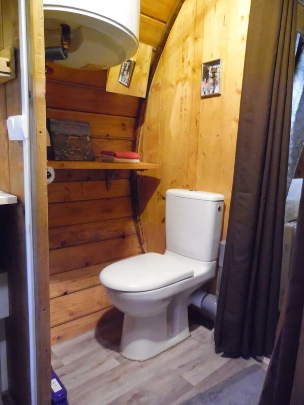 A small, wooden cabin-style bathroom with a white toilet, wooden shelves, and a shower curtain.
