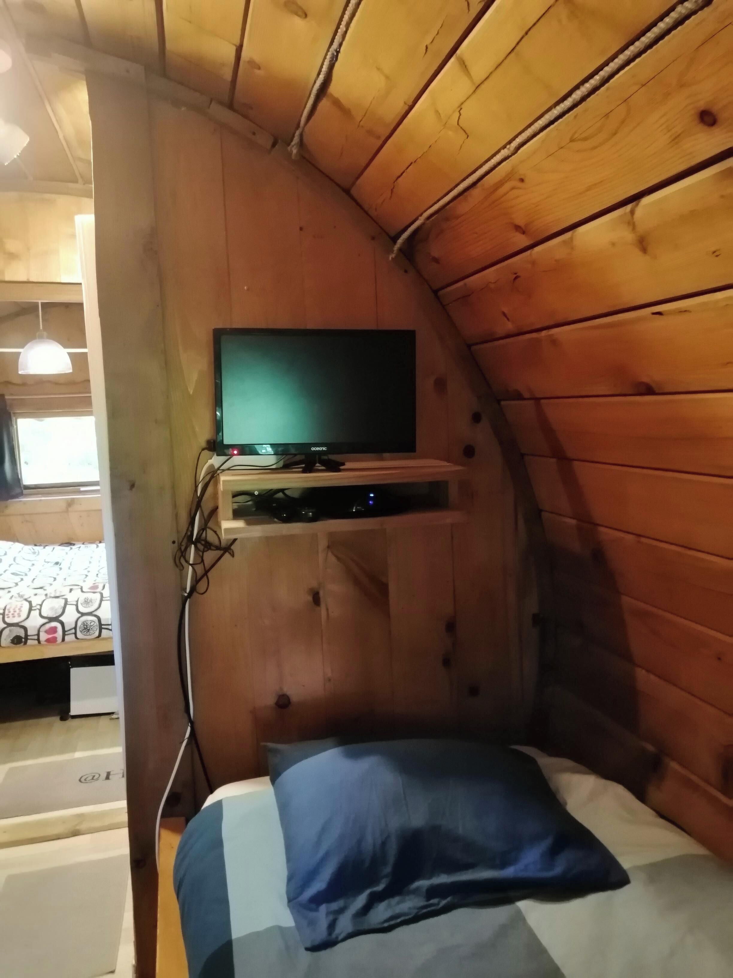 Ein kleiner, gemütlicher Raum mit Holzwänden, einem Bett mit blauem Kissen und einem angebrachten Fernseher.