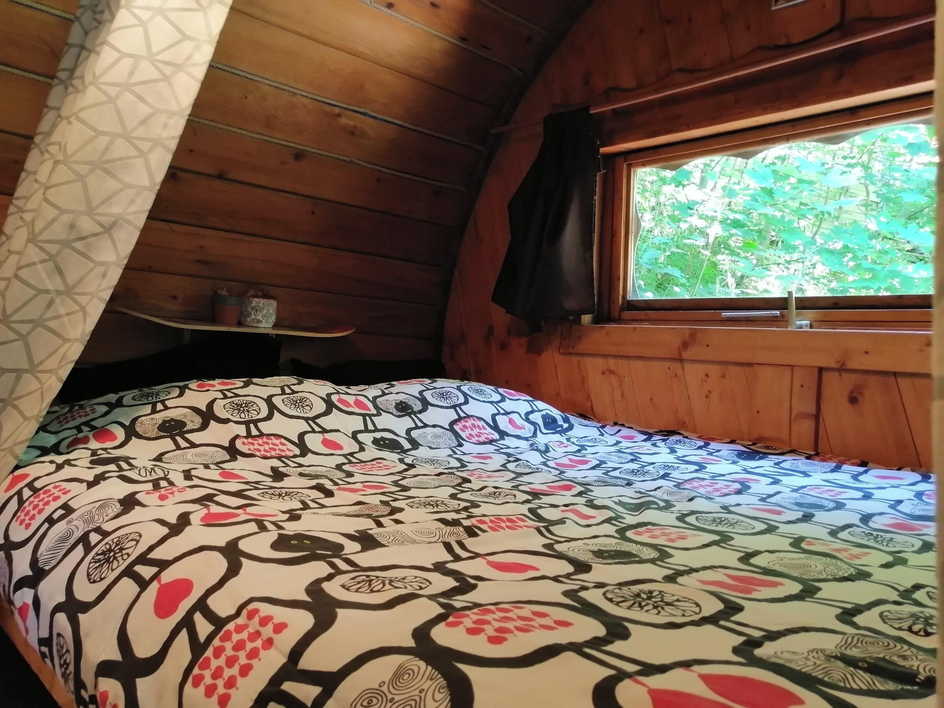A cozy room with a wooden sloped ceiling, a bed with a patterned bedspread, a window with a view of greenery, and a shelf with items.