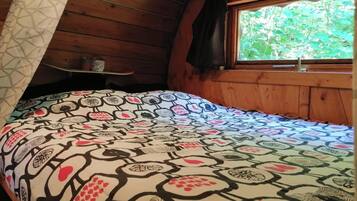 A cozy room with a wooden sloped ceiling, a bed with a patterned bedspread, a window with a view of greenery, and a shelf with items.