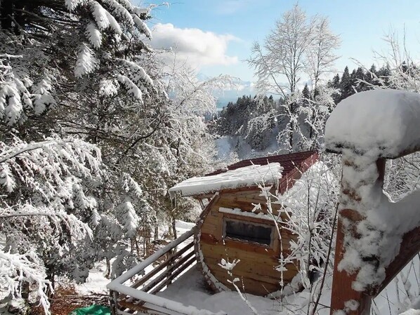 Une cabane en bois, dont le toit est recouvert de neige, entourée d’arbres chargés de neige et sous un ciel dégagé.