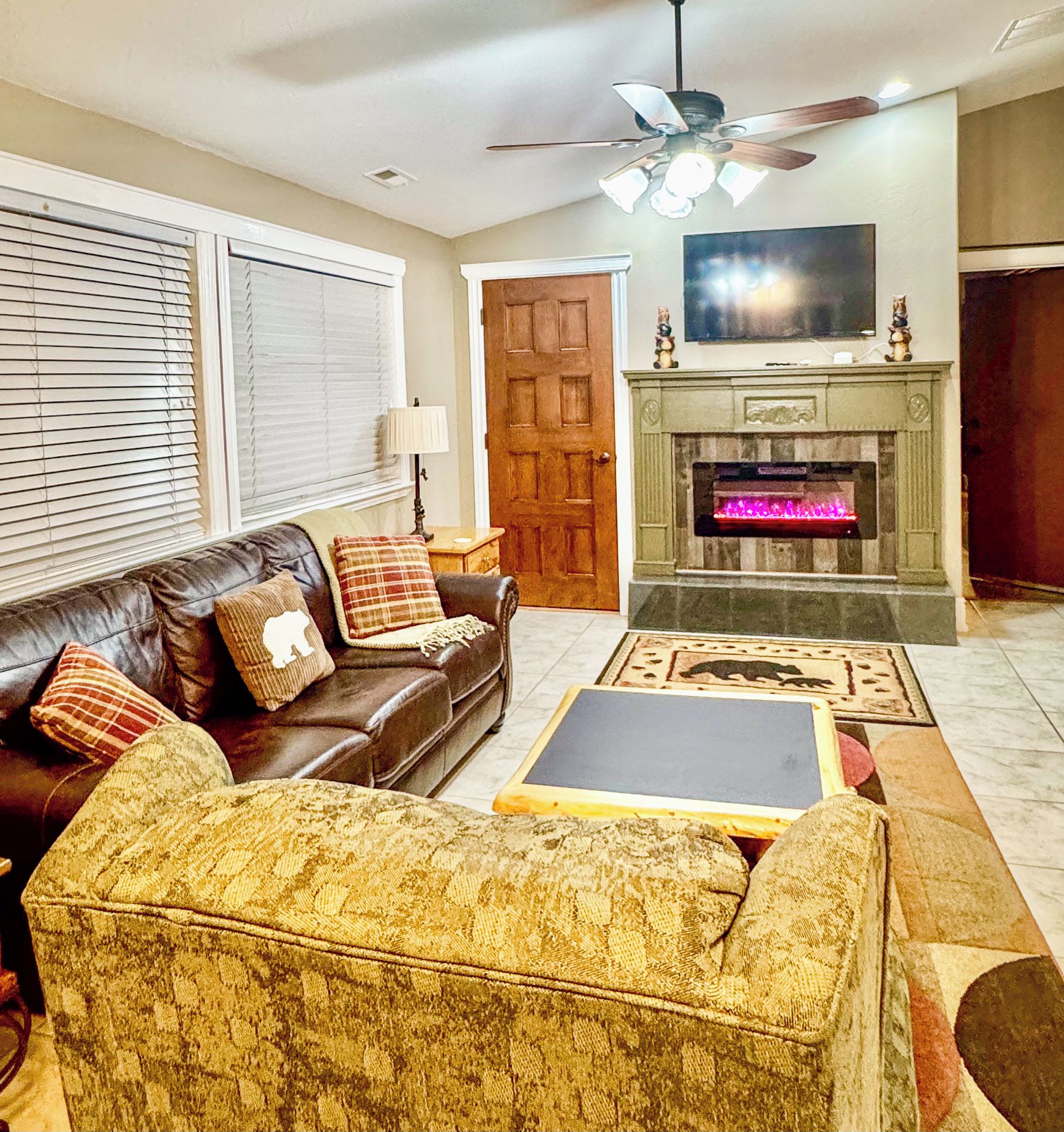 A living room with a leather sofa, patterned armchair, a fireplace with a lit fire, a ceiling fan, and a television on the wall.