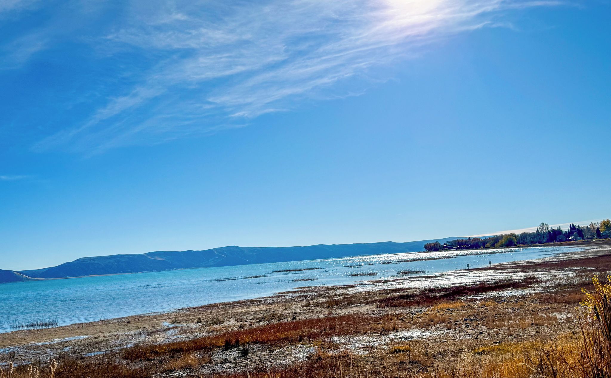A clear sky over a lake with distant mountains and a flat, grassy shoreline.