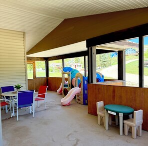 Een overdekt terras met een speeltoestel, buitenmeubels en uitzicht op een woonwijk.