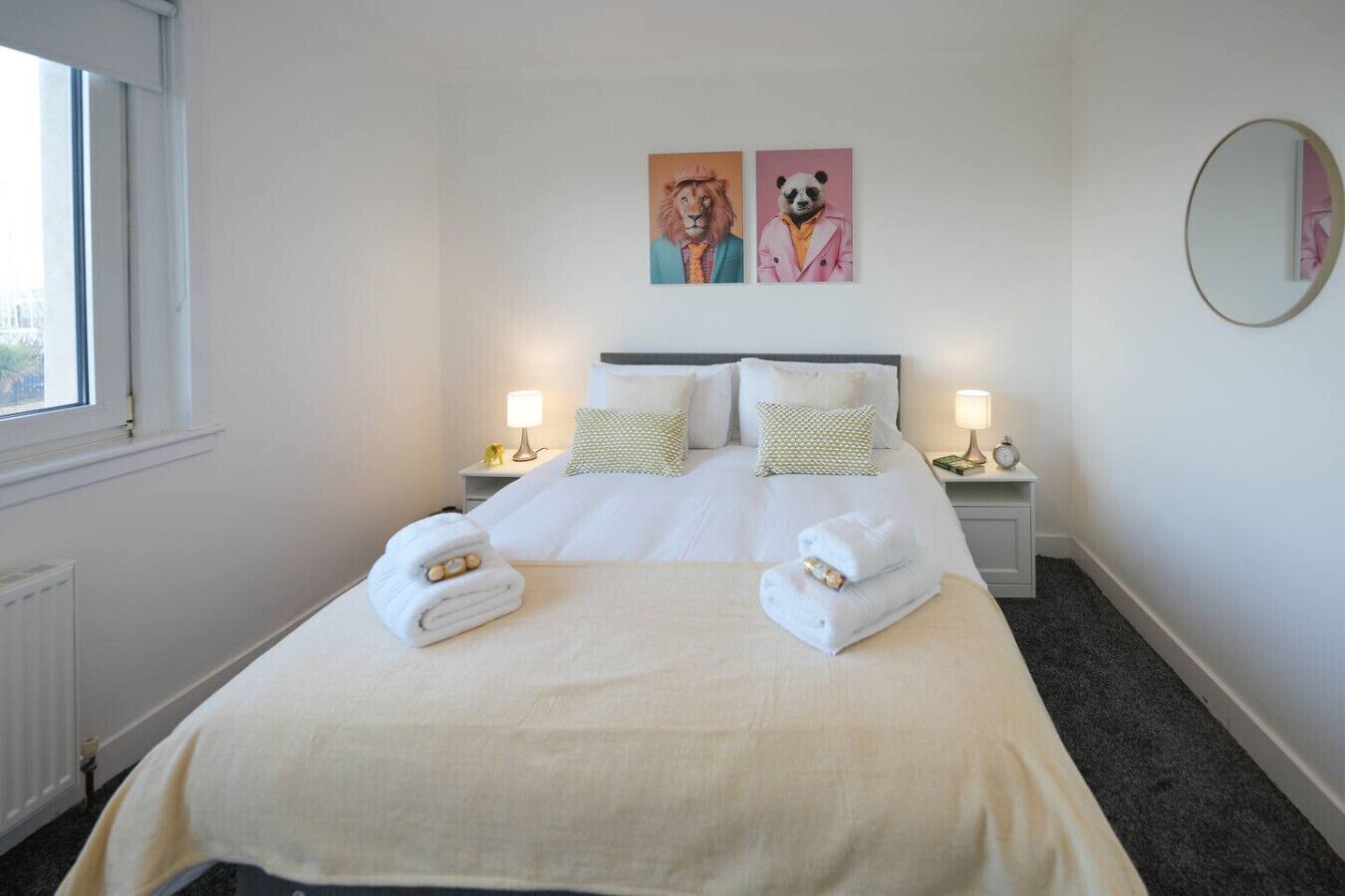 Een hotelkamer met een groot bed, twee nachtkastjes met lampen, een raam en twee dieren schilderijen aan de muur.