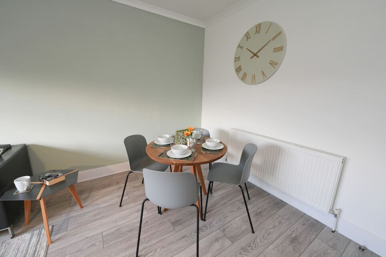 En spiseplads med et rundt bord dækket til fire personer, omgivet af grå stole, et hvidt vægur og en radiator.