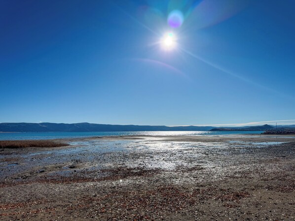 A beach with pebbles and water, mountains in the distance, and a clear sky.