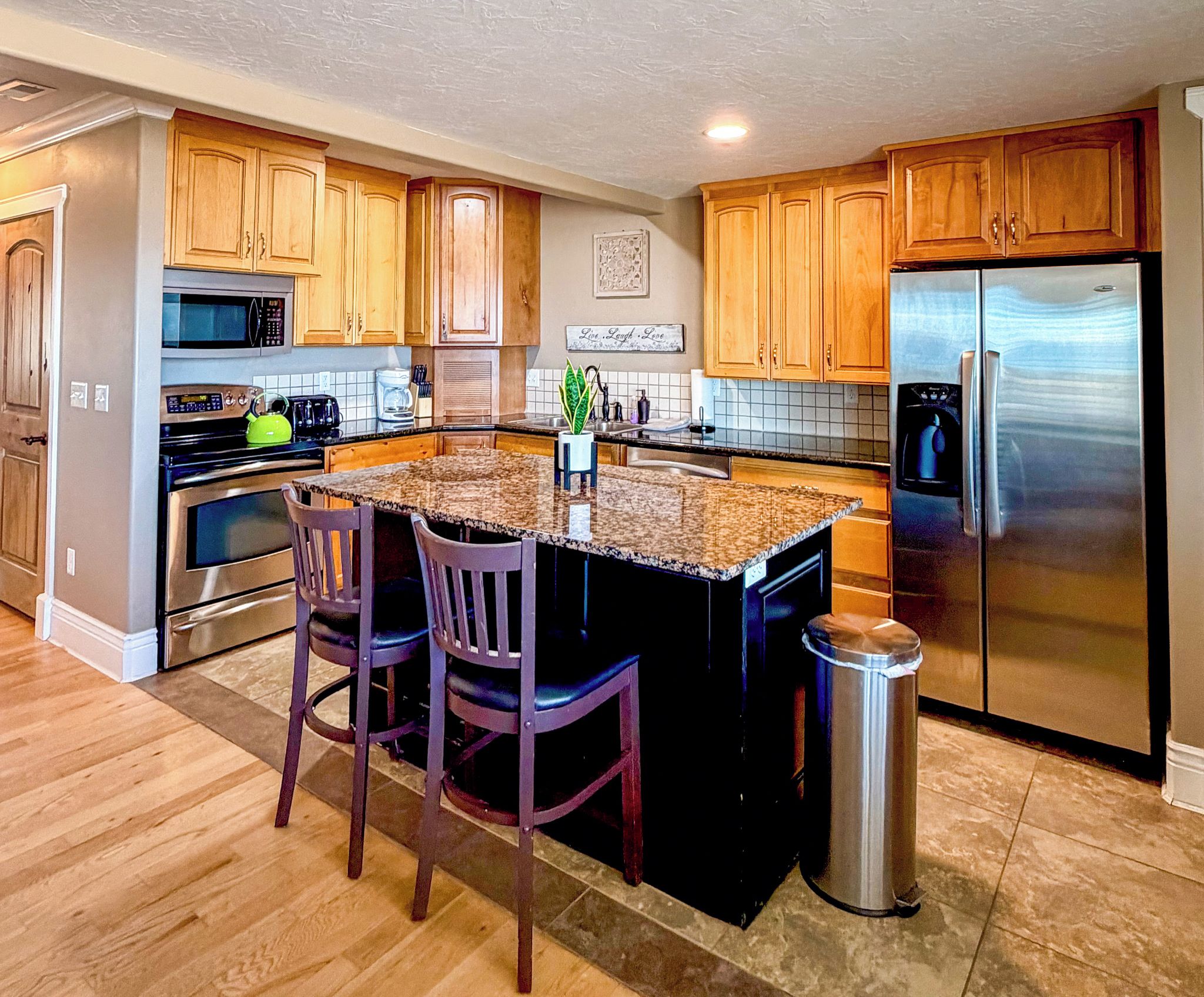 A kitchen with wooden cabinets, a stainless steel refrigerator, a microwave, and a granite countertop island with two chairs.
