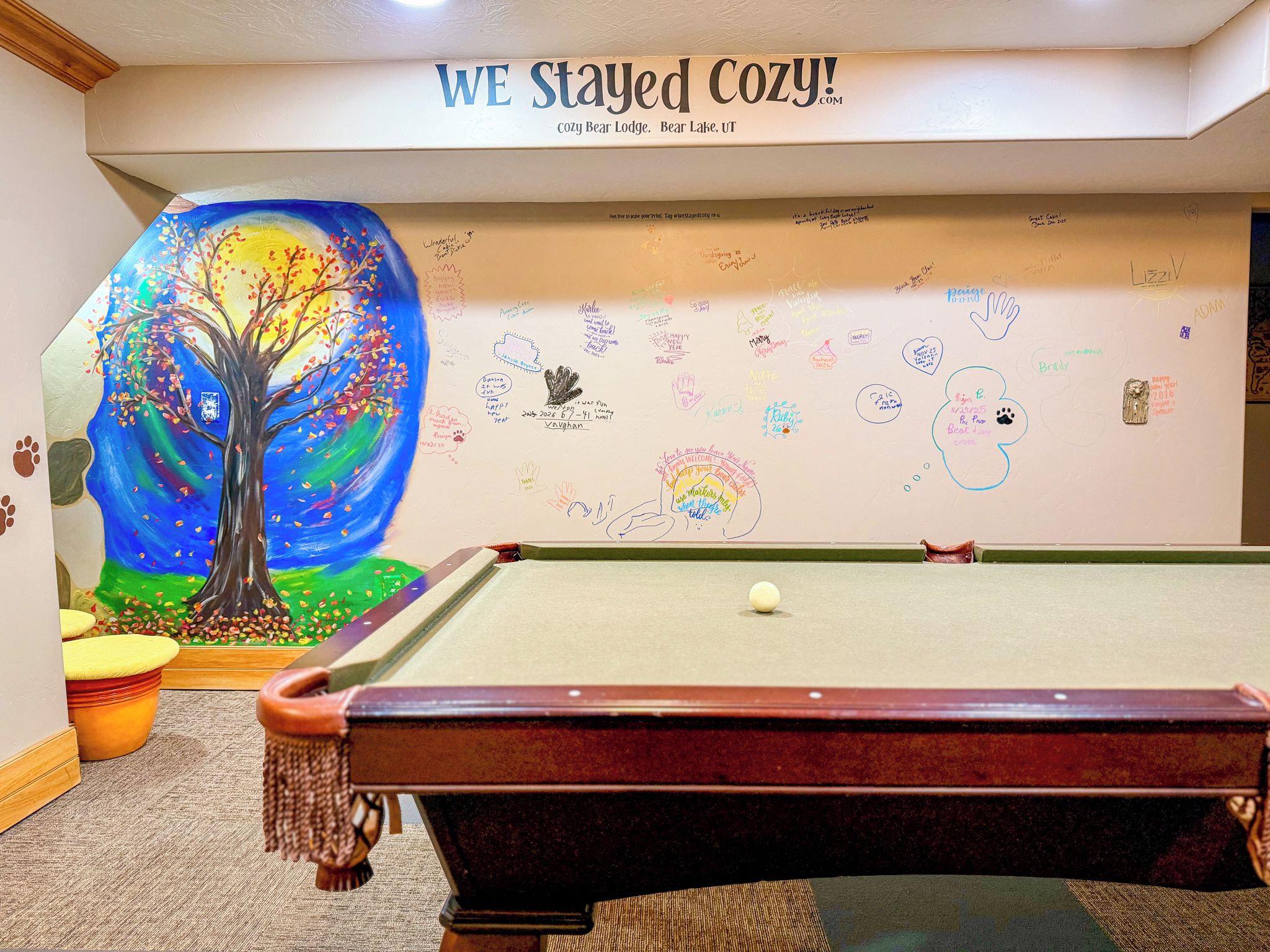 Une pièce chaleureuse avec une table de billard, une fresque représentant un arbre et un mur recouvert de divers messages et notes manuscrits.