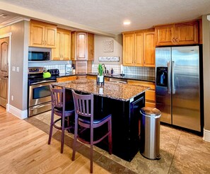 A kitchen with wooden cabinets, a stainless steel refrigerator, a microwave, and a granite countertop island with two chairs.