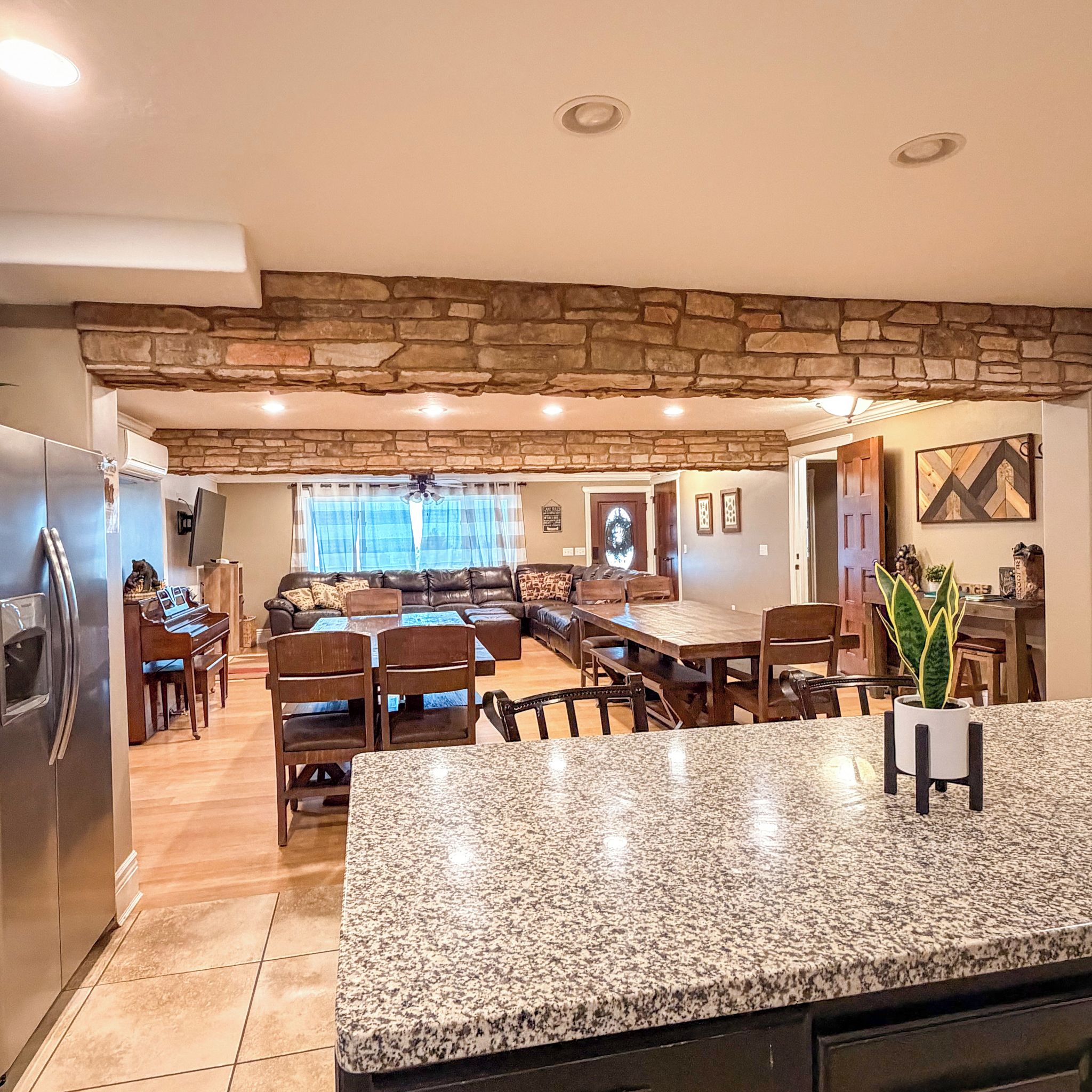 A spacious kitchen with granite countertops, a dining area with a wooden table and chairs, a stone accent wall, and a living room with a sofa and a fireplace.