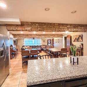 A spacious kitchen with granite countertops, a dining area with a wooden table and chairs, a stone accent wall, and a living room with a sofa and a fireplace.