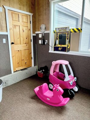 A room with a wooden door, a window with blinds, a Pac-Man arcade game, and a pink toy car.